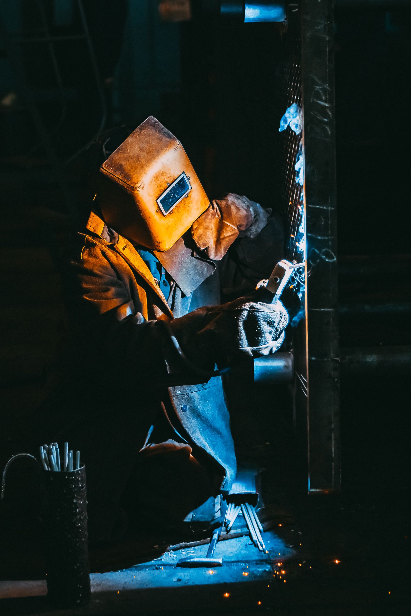 A welder in a full leather welding jacket and helmet, kneeling to weld a metal frame, with blue-white arc glow and sparks visible
